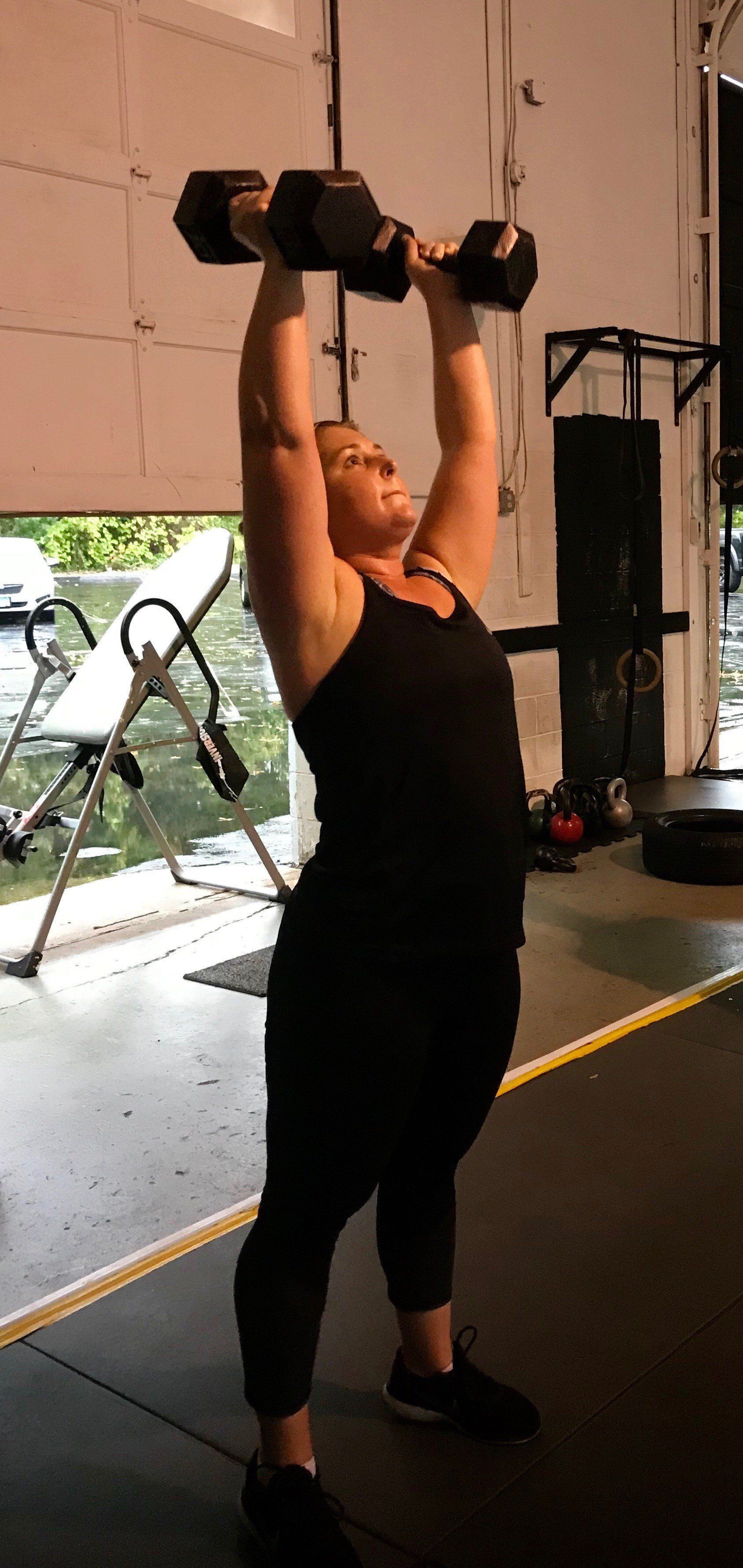 A woman is lifting dumbbells over her head in a gym.