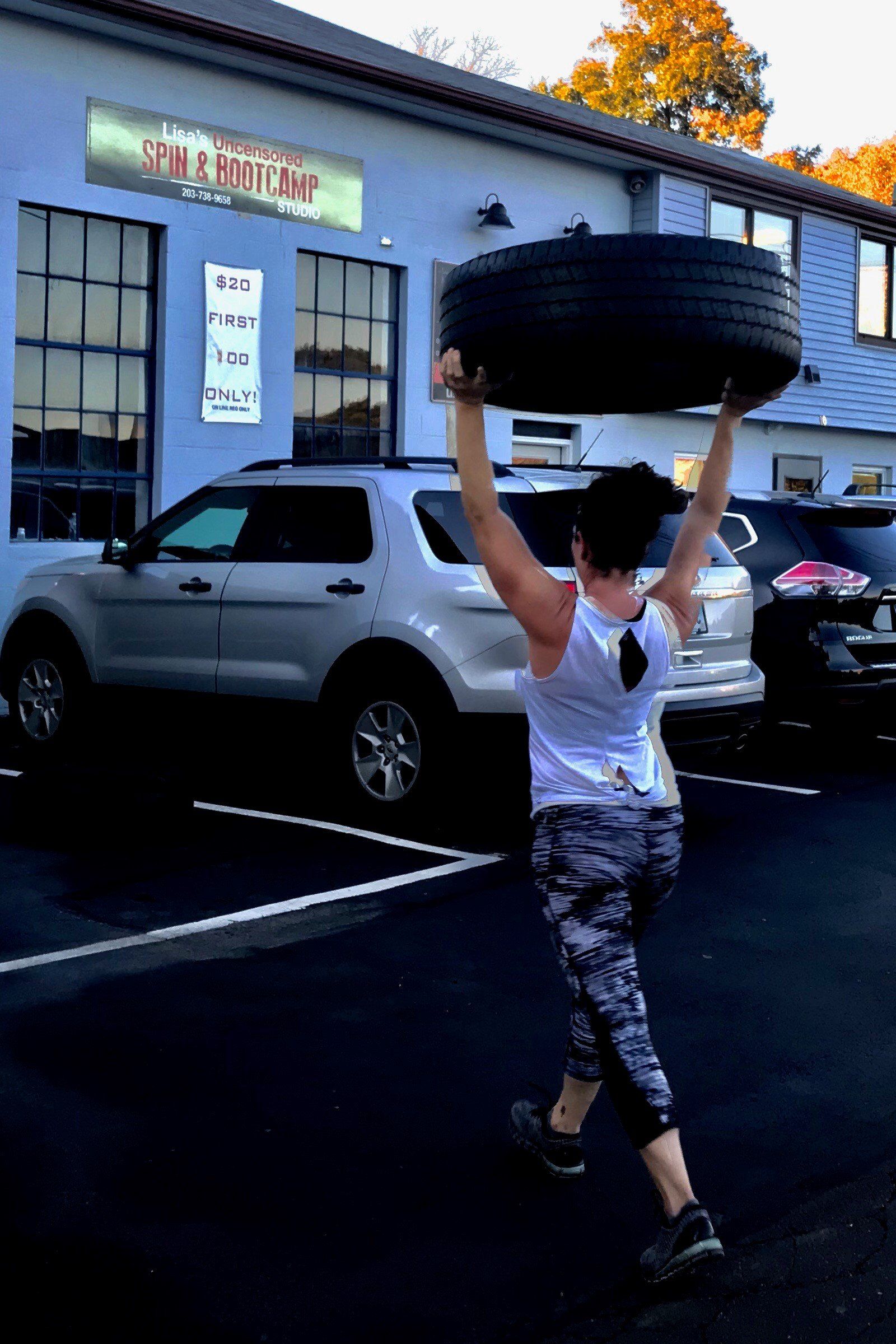 A woman is carrying a tire over her head in a parking lot.