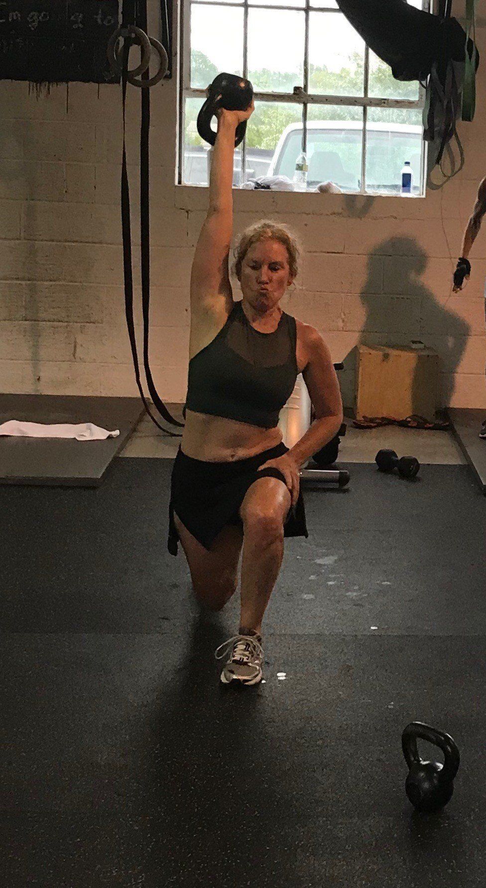 A woman is squatting down in a gym while holding a kettlebell over her head.