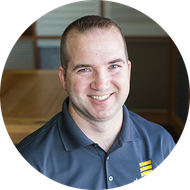 Man with short brown hair smiles, wearing a gray polo shirt, set in an office.