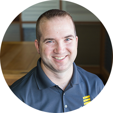 Man with short brown hair smiles, wearing a gray polo shirt, set in an office.