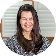 Woman with long brown hair smiling, wearing a gray and white patterned sweater in front of white shutters.