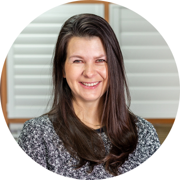 Woman with long brown hair smiling, wearing a gray and white patterned sweater in front of white shutters.
