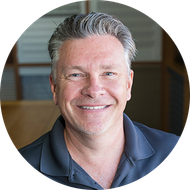Smiling man with graying hair in a dark polo shirt, indoors, slightly blurred background.