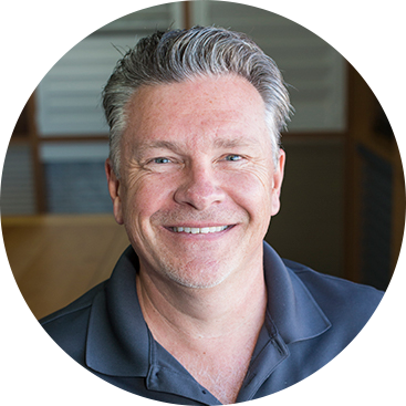 Smiling man with graying hair in a dark polo shirt, indoors, slightly blurred background.