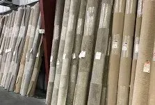 Rolled-up carpet rolls of various colors and textures stand vertically in a store or warehouse setting.
