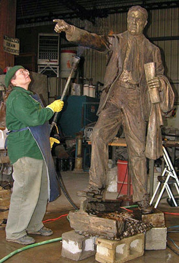 A person in a green shirt and apron uses a handheld torch to work on a bronze statue of a man gesturing forward.