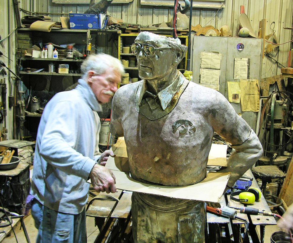Artist working beside a large bust sculpture in a cluttered workshop, surrounded by tools and shelves