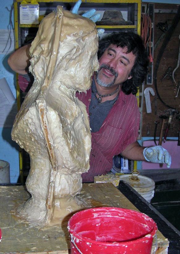 A person with a mustache creates a sculpture in a workshop, applying a thick, light-brown material to a bust.