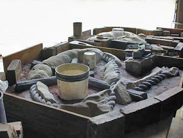 A sandy sand-casting mold in a wooden frame with a bucket, stone tools, and circular impressions for metal casting.