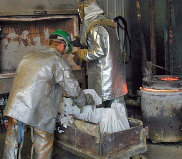 Two workers in reflective heat-protective suits and safety gear prepare to pour molten metal from a furnace.