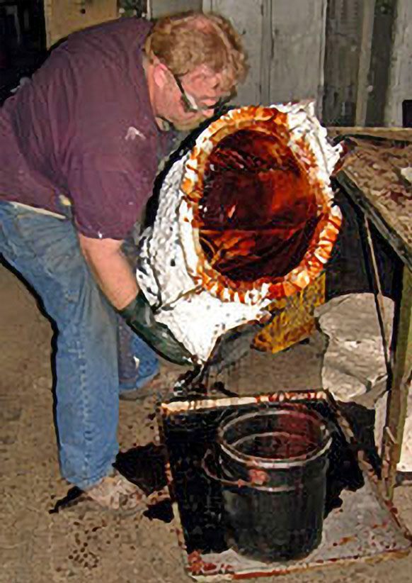 A person wearing protective gloves tilts a large, coated, industrial-looking mold to pour thick, dark liquid into a bucket.