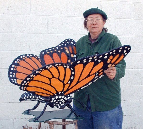 A person in a dark green shirt and beret holds a large, brightly painted monarch butterfly sculpture.