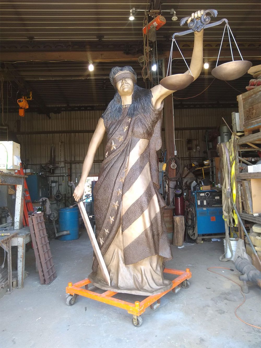 A statue of Lady Justice, blindfolded and holding scales and a sword, stands on a wheeled dolly inside a workshop.