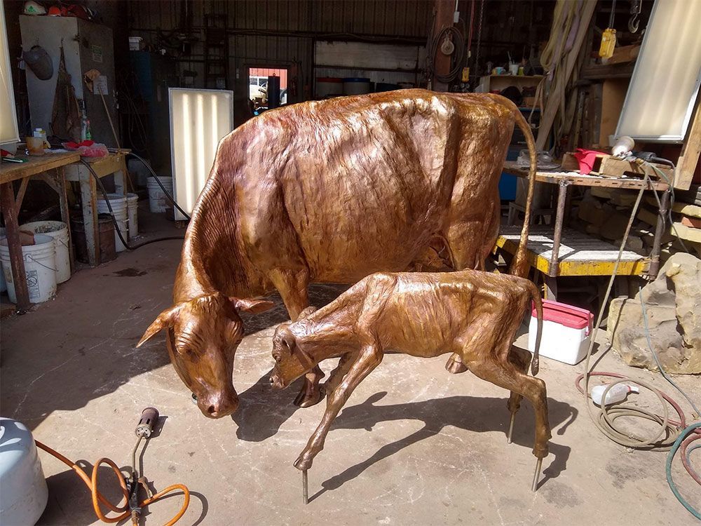 A copper-colored metal sculpture of a cow and its calf standing inside a workshop.