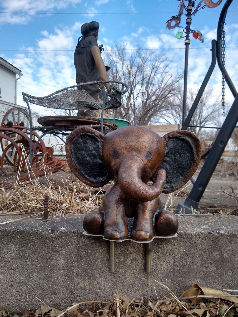 A bronze-colored, cartoon-style elephant garden stake sits on a concrete ledge with metal garden art in the background.
