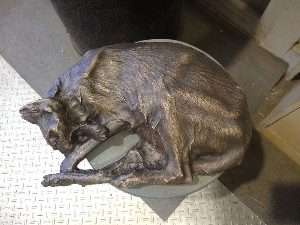 A bronze sculpture of a thin, curled-up dog resting on a circular base with a textured metallic floor in the background.