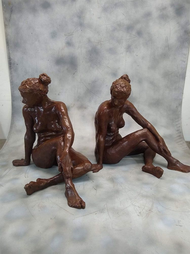 Two brown, seated clay sculptures of human figures posing on a mottled gray background.