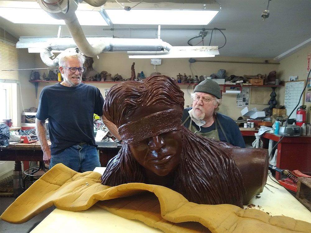 Two people stand behind a sculpture of a figure wearing a headband in a workshop with tools and supplies.