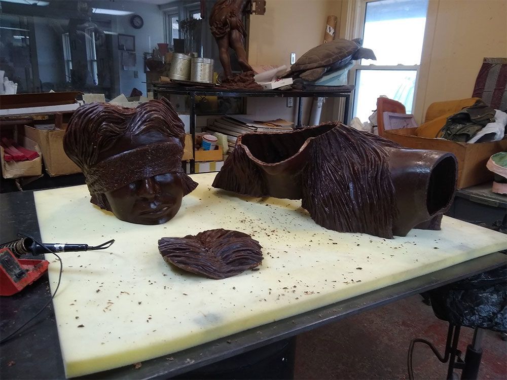 Sculpted dark chocolate pieces, including a bust and a textured form, sit on a foam pad in a workspace.
