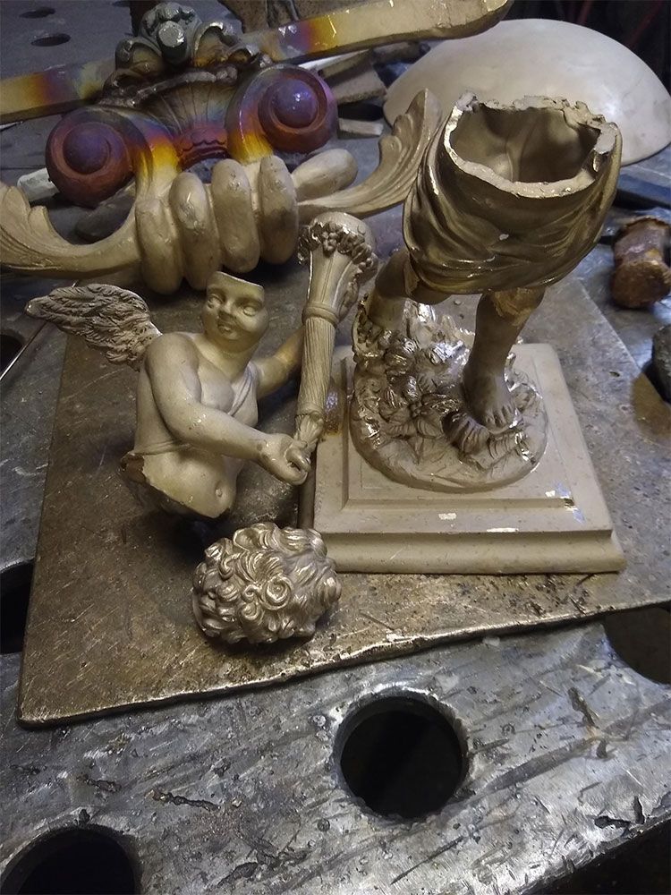 Broken cream-colored statue pieces, including a cherub and torso, resting on a workshop metal surface.