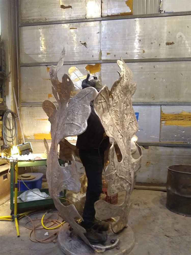 A person stands inside a tall, cylindrical metal sculpture shaped like large, textured oak leaves in a workshop.
