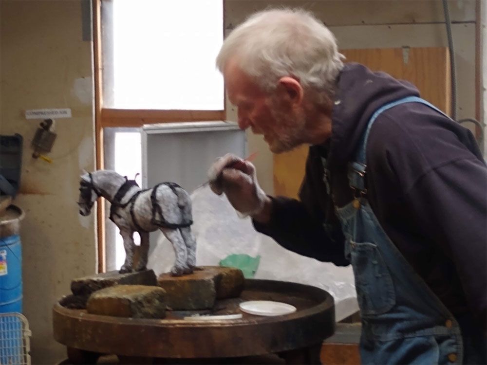 A person wearing overalls carefully paints a detailed model of a horse in a workshop setting.