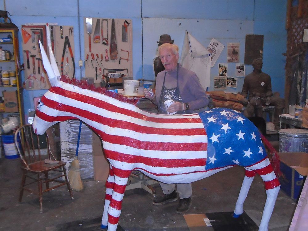 An artist works in a studio on a life-sized donkey sculpture painted with red and white stripes and a blue star field.