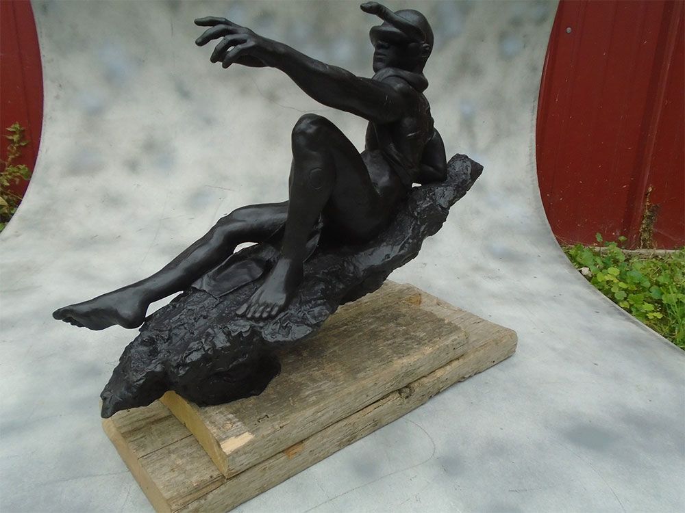 A black sculpture of a seated figure wearing a winged helmet, reaching forward, set on a textured base and wooden plank.