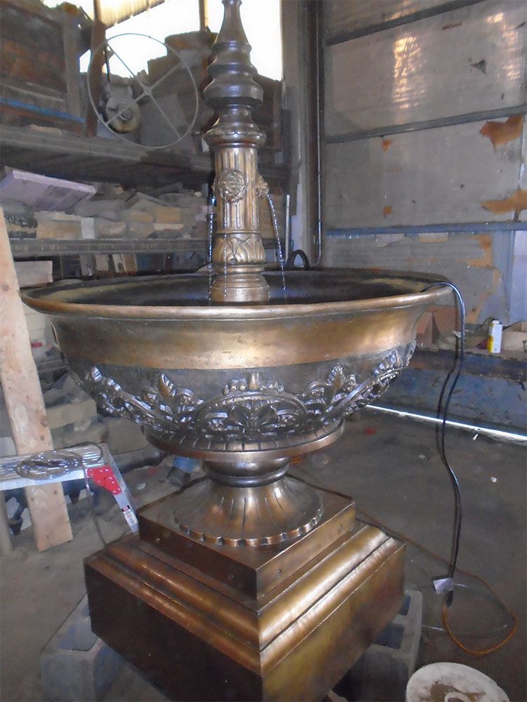 A large, ornate, metallic fountain with a central spire, sitting on a square base in a workshop setting.