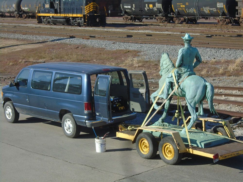 A blue van tows a trailer carrying a green bronze equestrian statue, parked near train tracks on a paved surface.
