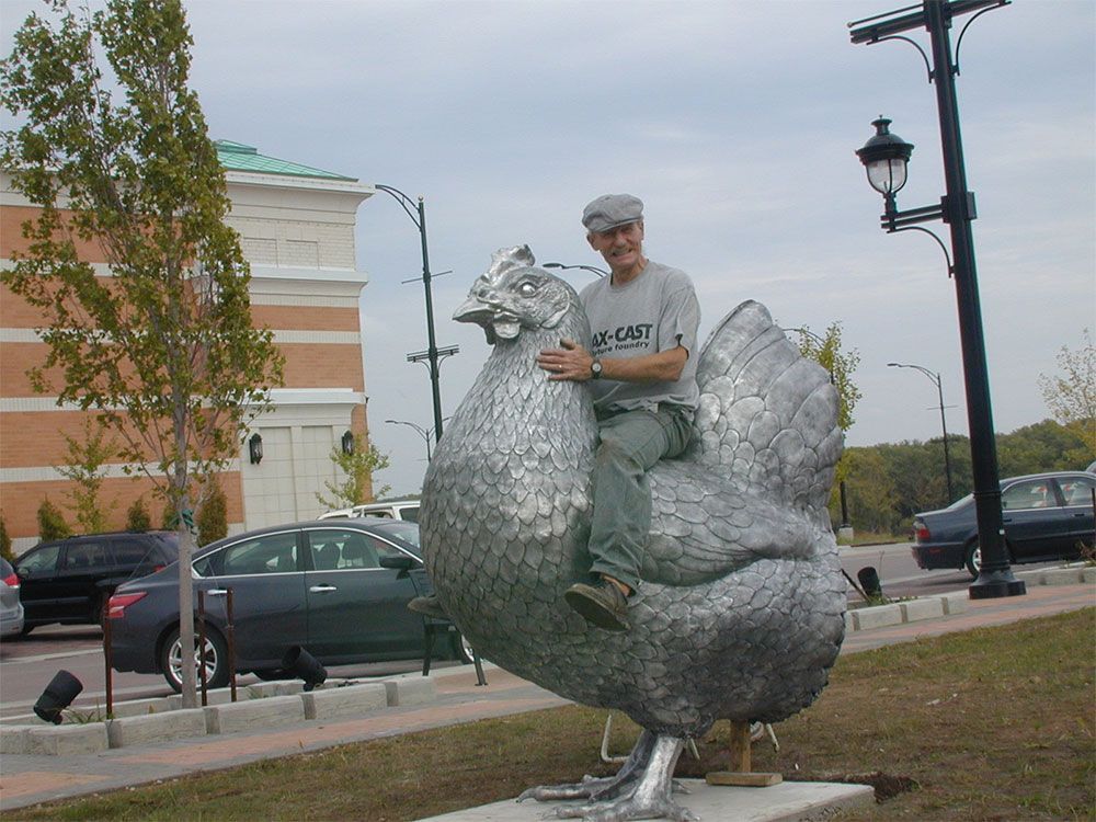 A person wearing a grey cap and t-shirt sits atop a large, metallic chicken sculpture in a parking lot setting.