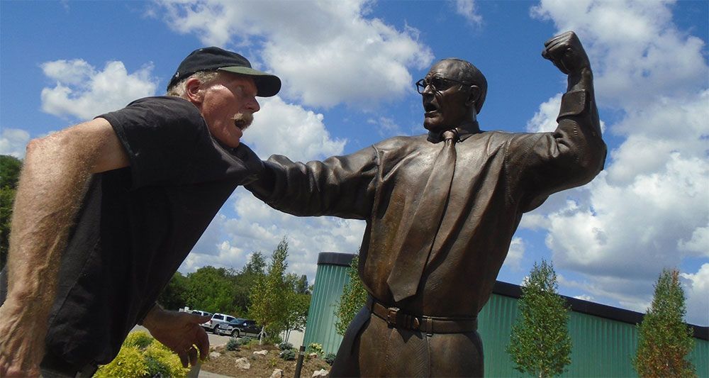A person leans back as if startled while standing next to a bronze statue of a person shouting with an arm raised.