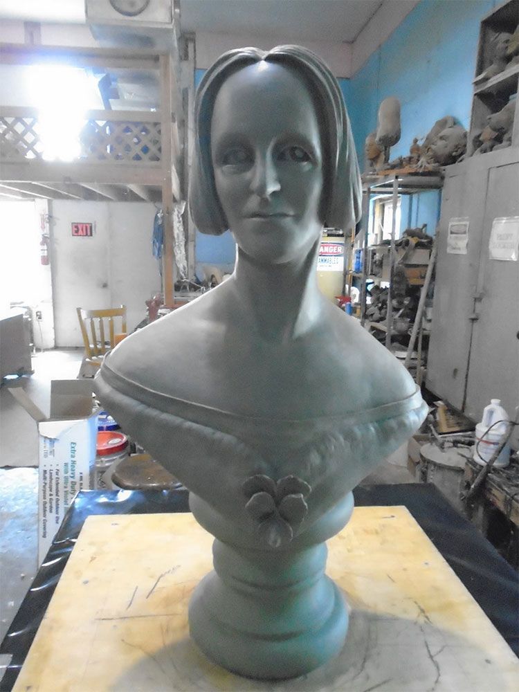 A gray clay sculpture of a person’s head and torso, wearing a garment with a floral detail, in an art studio.