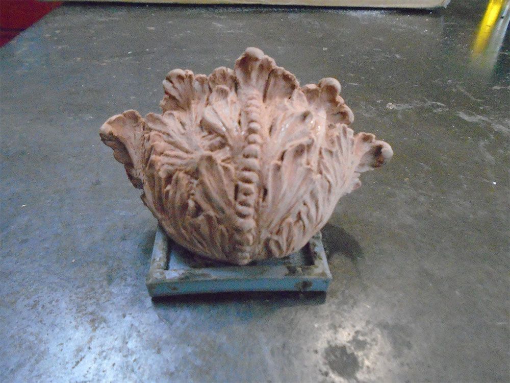 A terracotta sculpture shaped like a leafy vegetable, resting on a small, square blue metal frame on a gray surface.