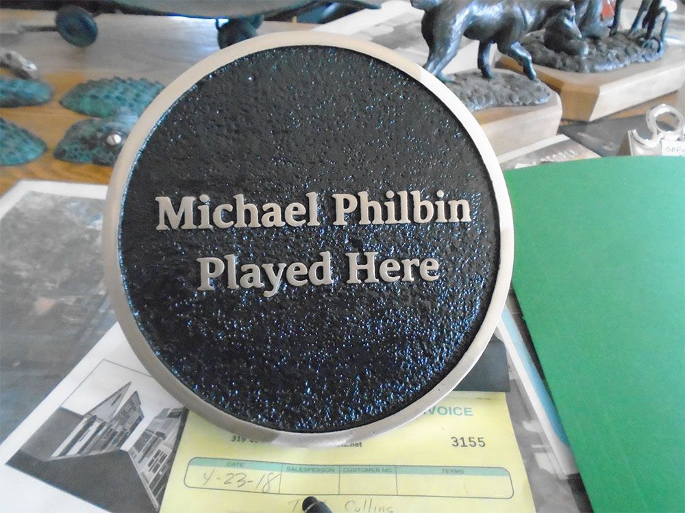 A circular, textured black memorial plaque with silver trim and the engraved text