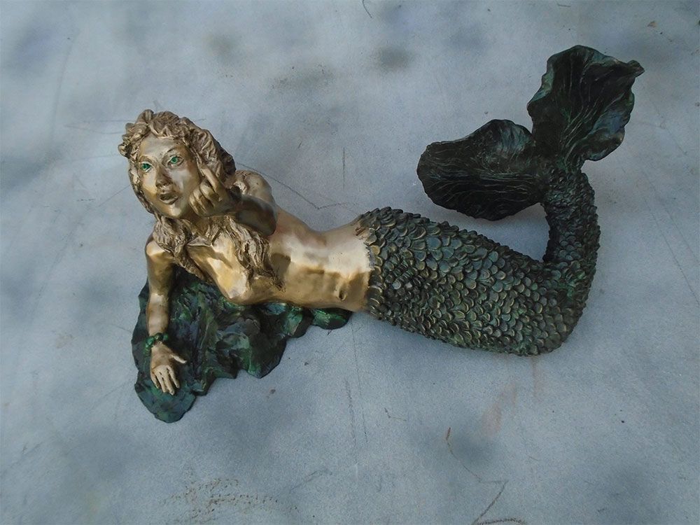 A bronze-toned mermaid statue resting on a rocky base, featuring textured scales and a prominent, flared tail.