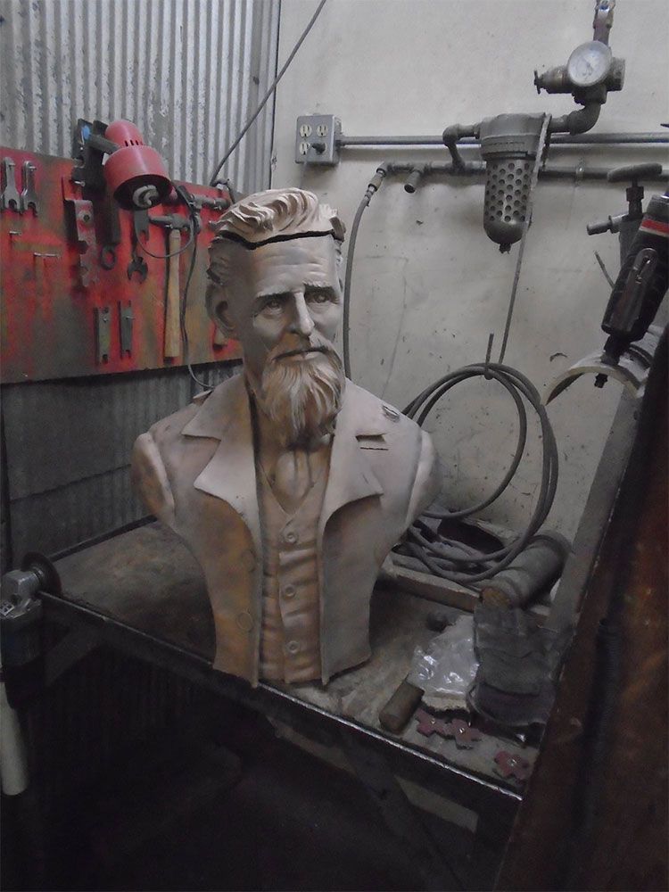A light-colored bust of a bearded man in a suit, placed on a workbench inside a cluttered workshop.