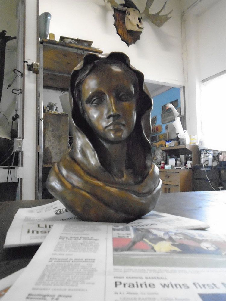 A bronze sculpture of a woman’s head draped in a shawl, sitting on a newspaper in a workshop with a moose mount above.