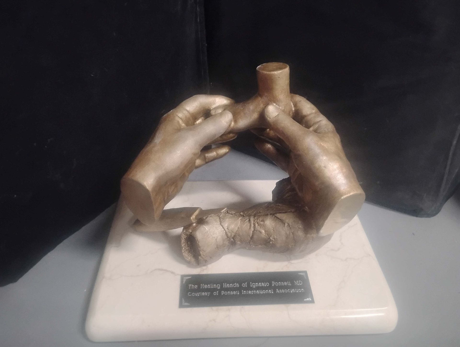 Bronze sculpture of two hands holding a small cup on a white pedestal with a plaque