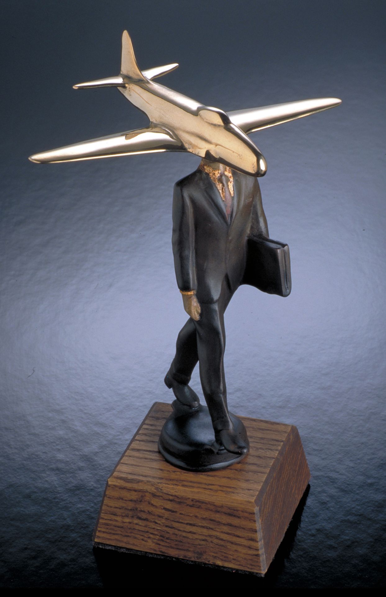 Surreal sculpture of a suited figure with a jet plane for a head, mounted on a wooden base.