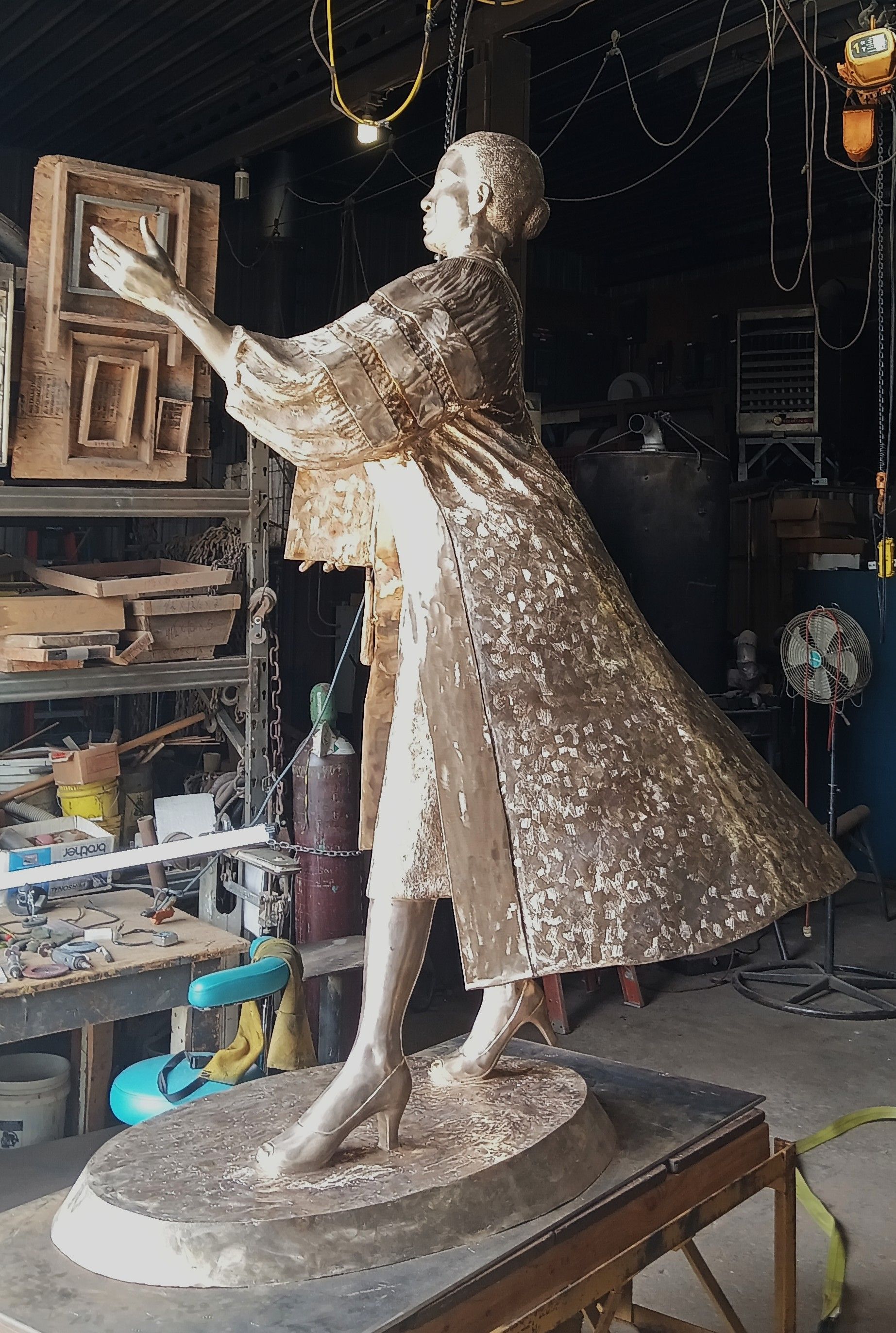 Bronze-colored statue of a woman in a flowing dress, displayed in a workshop studio.