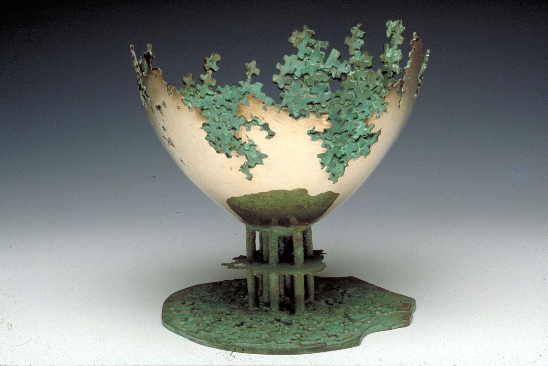 A green leafy sculpture shaped like a bowl on a stand, set on a grassy base against a plain background