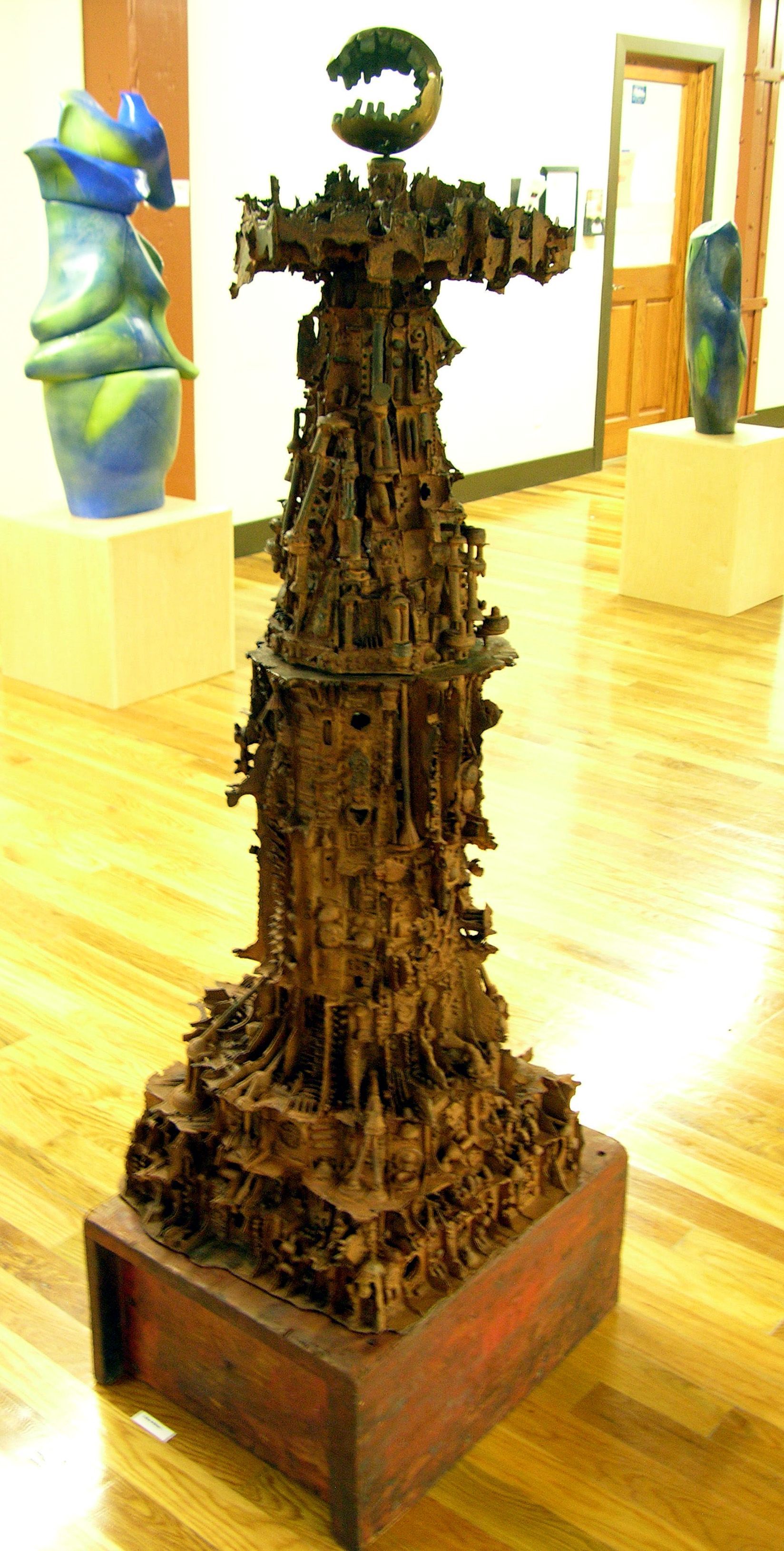 Small dark carved sculpture on a wooden base in a bright gallery room, with colorful art in the background