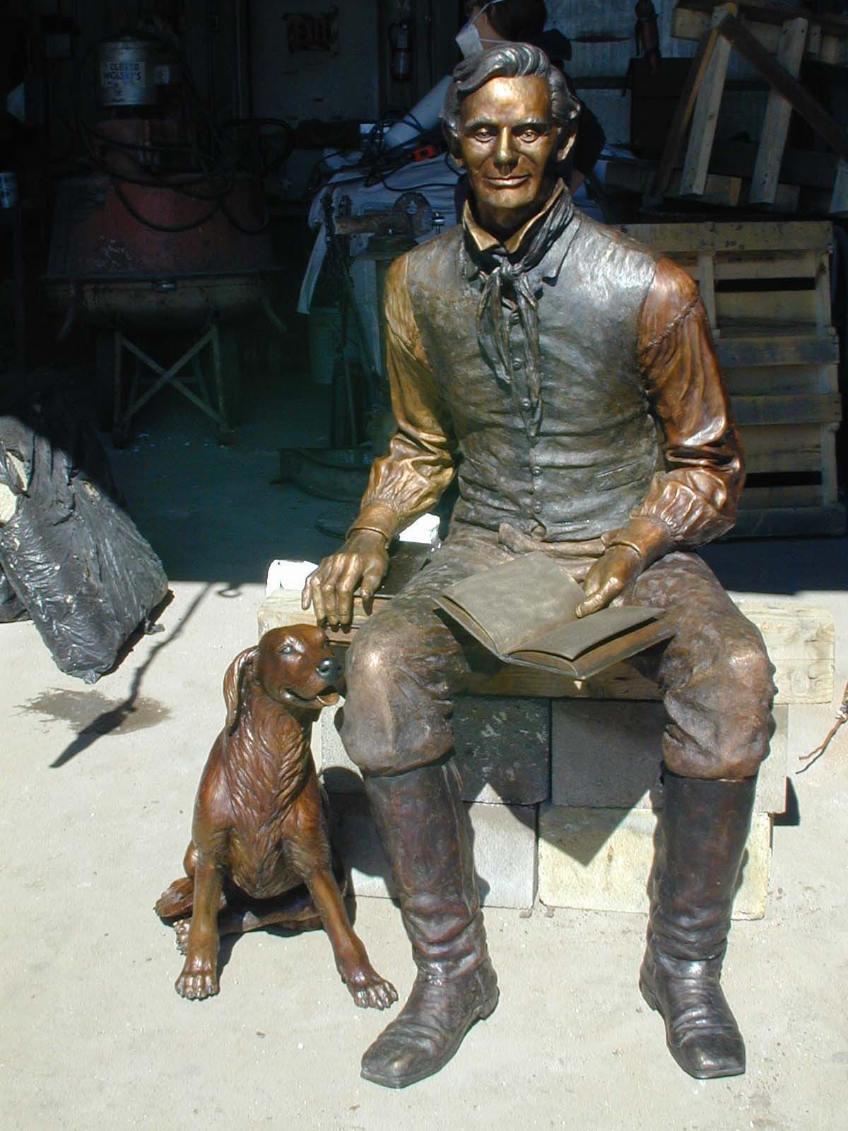 Statue of a seated man in rustic clothes with a dog at his feet, displayed outdoors.