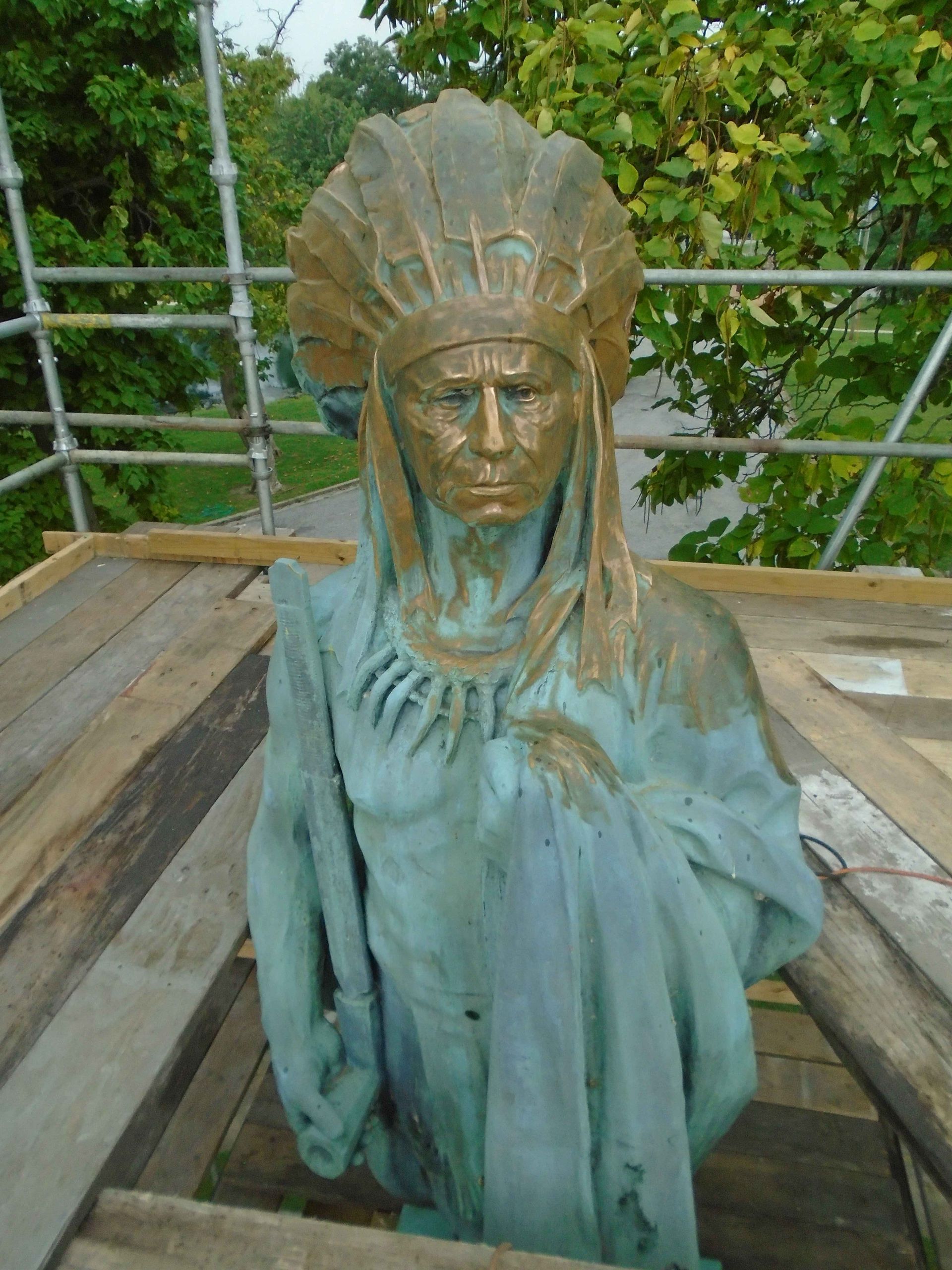 Weathered green statue bust with feathered headdress on a wooden platform outdoors