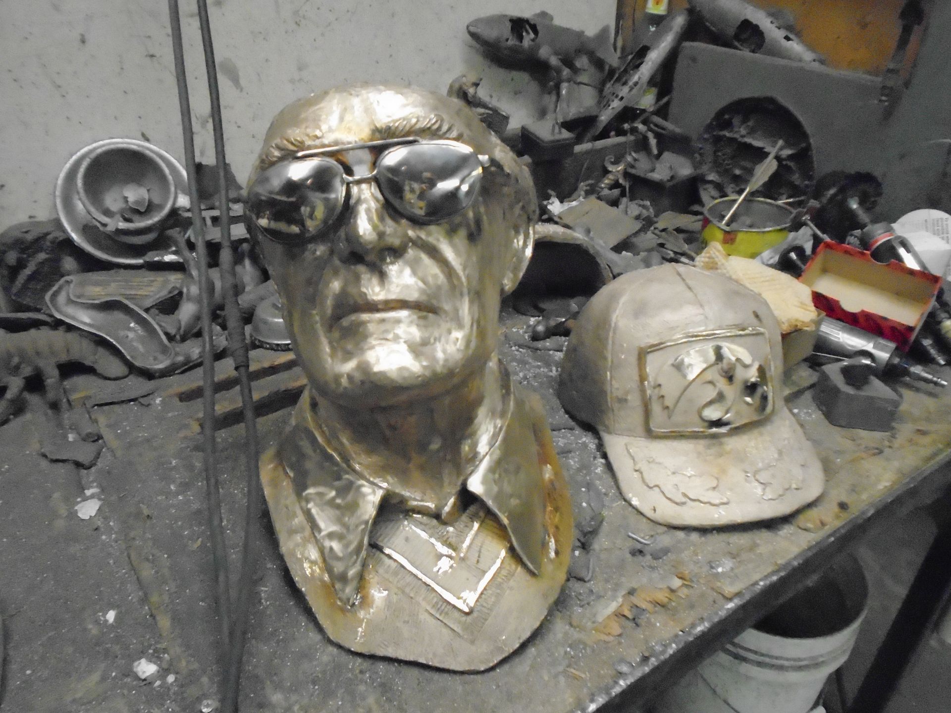 Gold bust and white cap displayed on a workbench with tools and metal parts in a cluttered workshop.