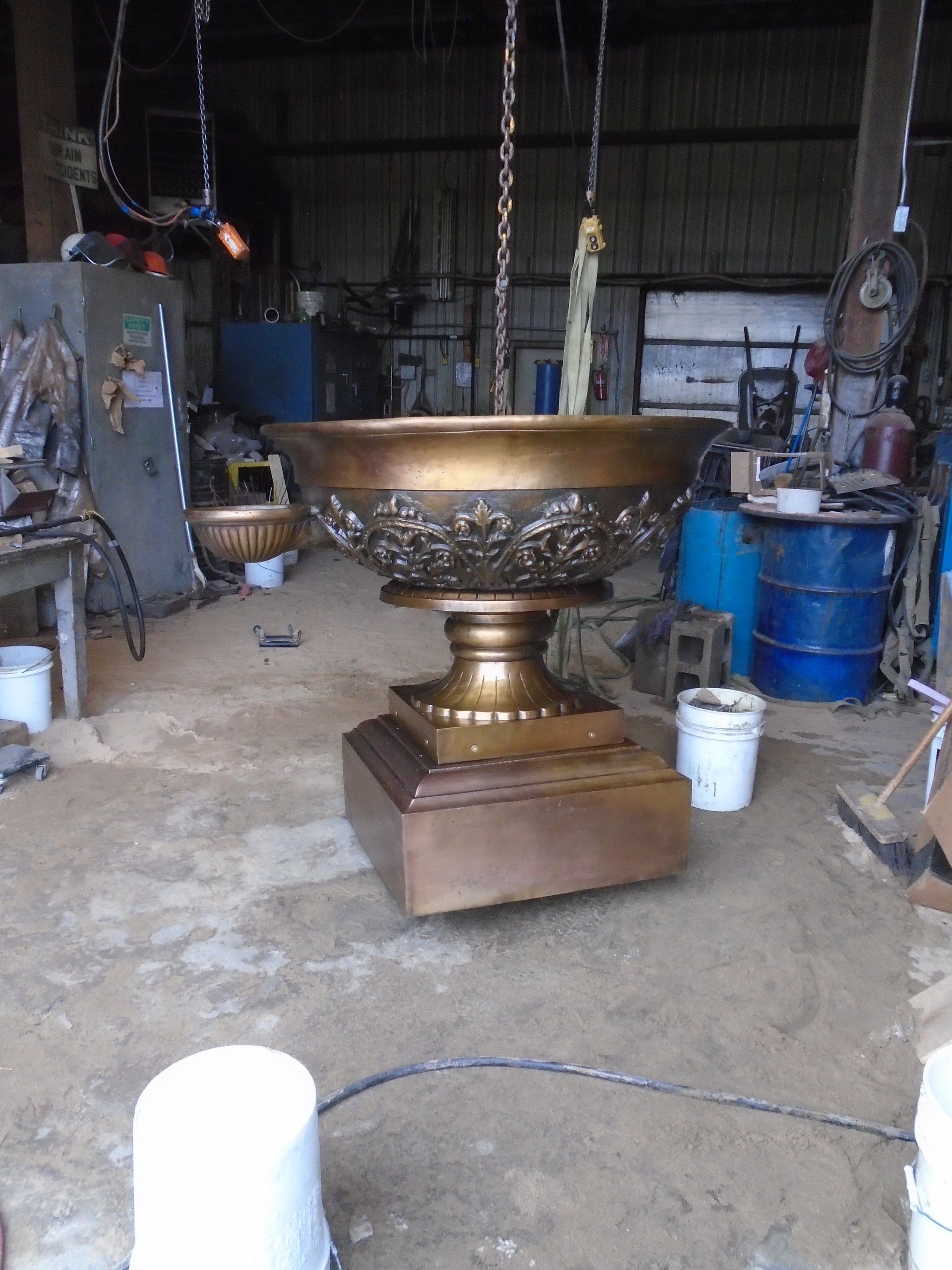 Large ornate gold bowl on a pedestal in a workshop, with buckets and tools around it.