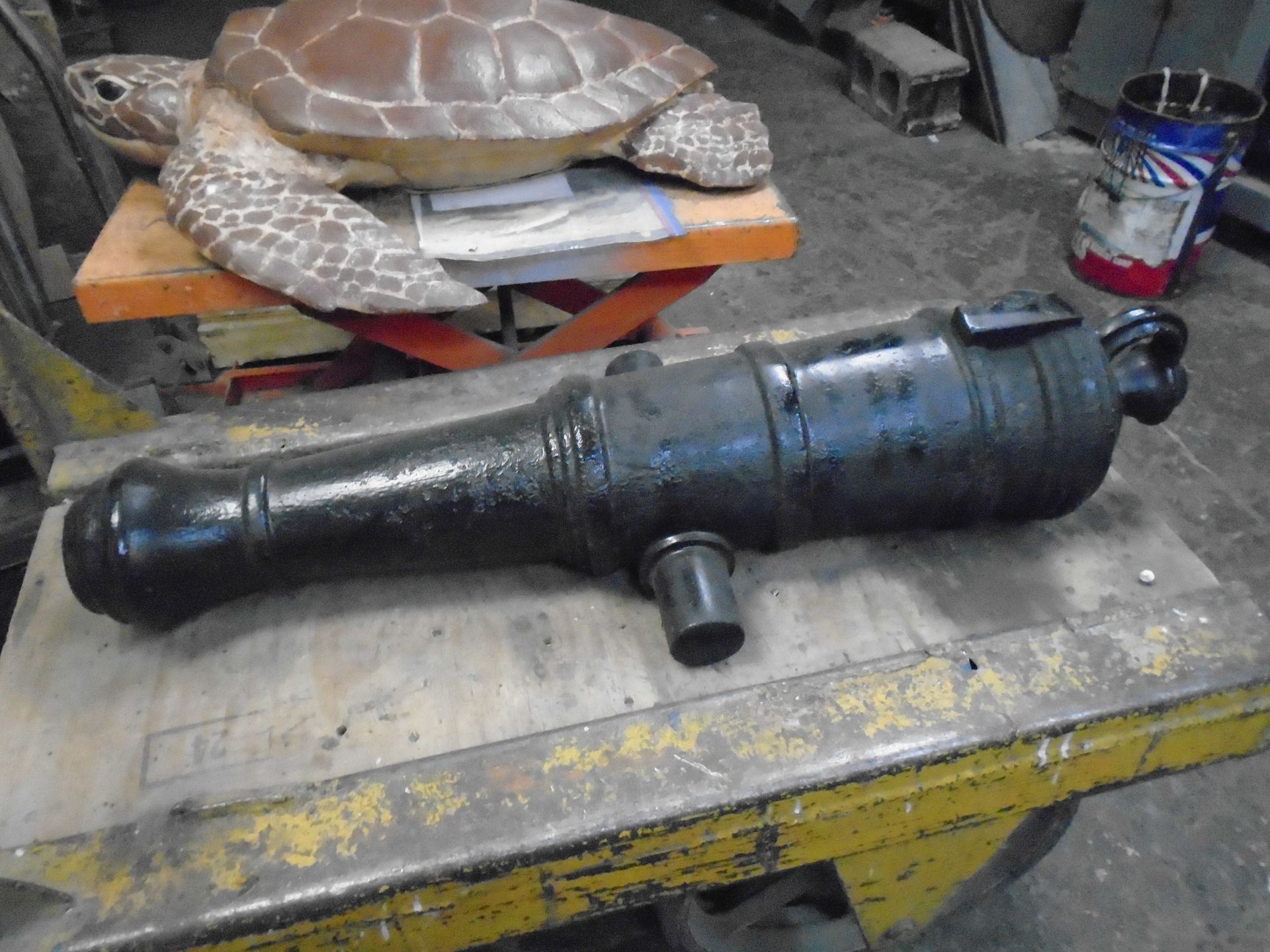 Black metal cylindrical machine part on a workbench in a workshop, with a turtle sculpture in the background
