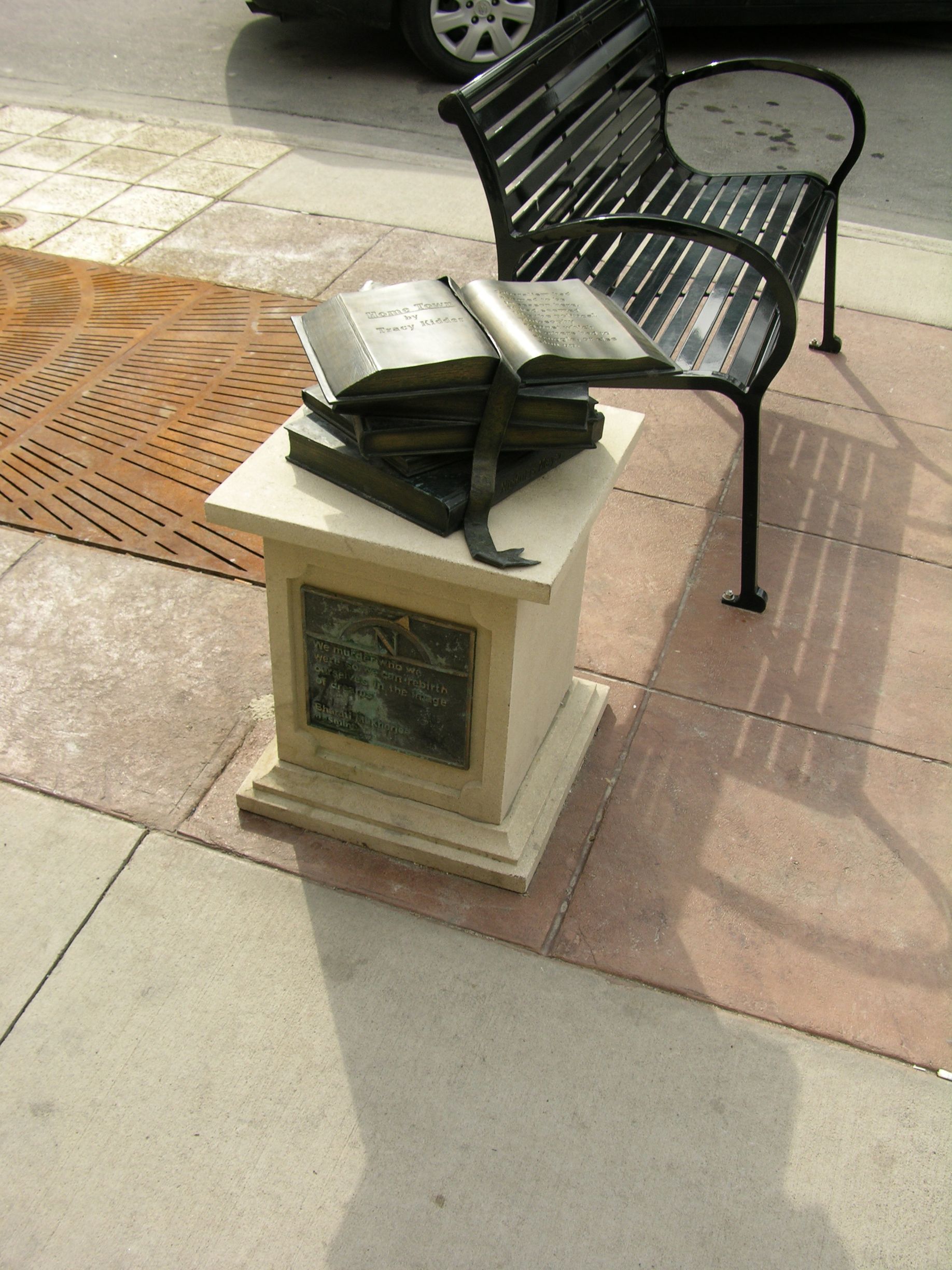 Open book on a small pedestal beside a black metal bench on a sidewalk.